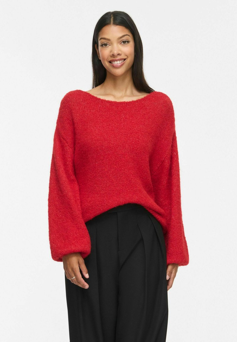 Femme aux longs cheveux noirs portant un pull rouge duveteux et un pantalon noir ample, souriant sur un fond blanc uni.