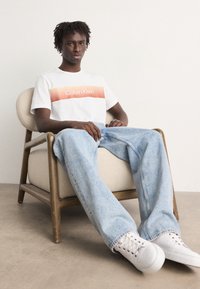 Bílá tričko s oranžovým gradientem loga „Calvin Klein“, v kombinaci se světle modrými širokými džínami a bílými teniskami, sedící na dřevěné židli.