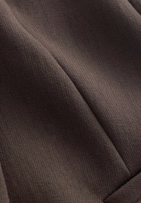 Detailní záběr tmavě hnědé texturované látky s viditelným švem a měkkými záhyby vytvářejícími stíny a hloubku.