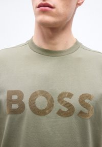 Άνδρας που φοράει χακί μπλουζάκι με στρογγυλή λαιμόκοψη και μεγάλα ανάγλυφα γράμματα που σχηματίζουν τη λέξη "BOSS" στο στήθος, εμφανιζόμενος από τον λαιμό έως το άνω μέρος του κορμού.