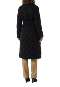 Black quilted long coat with a belted waist, featuring diamond-patterned stitching and a smooth texture. Paired with beige trousers and black heels.