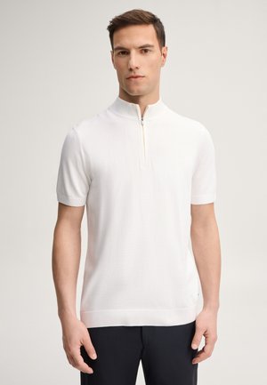 Uomo in piedi in una stanza semplice, indossa una maglia bianca a maniche corte in maglia con collo a mezza zip e pantaloni neri, rivolto in avanti, mani rilassate.