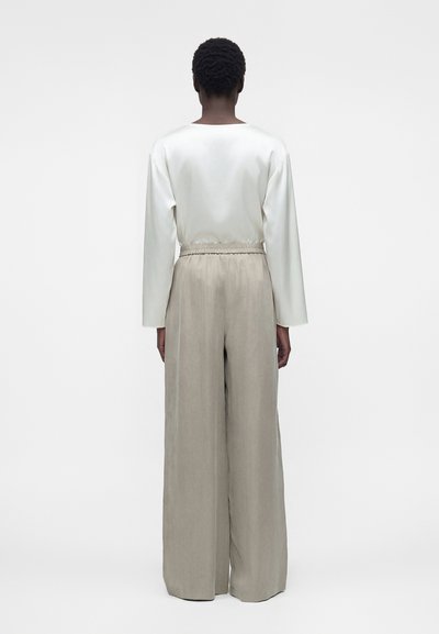 Άτομο με κοντά σγουρά μαλλιά που φοράει λευκό τοπ με μακριά μανίκια και μπεζ παντελόνες με ψηλή μέση και φαρδιά μπατζάκια, στέκεται με την πλάτη στραμμένη.