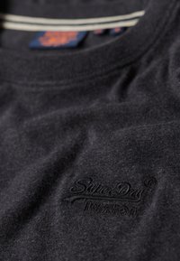 Donkergrijze stof close-up met een geborduurd logo met sierlijk schrift "SuperDry" en kleinere tekst eronder op het borstgedeelte van een kledingstuk.