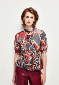 Blusa colorata e stampata con colletto a fiocco, maniche corte e voluminose, e vita a peplum, realizzata in tessuto leggero. Abbinata a pantaloni in pelle rossa.