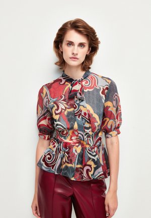 Blusa colorida y estampada con cuello de lazo, mangas cortas y abullonadas, y cintura peplum, fabricada en tejido ligero. Combinada con pantalones de cuero rojos.