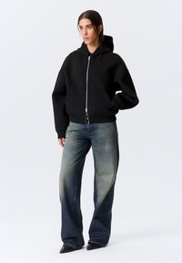 Kobieta ubrana w czarną bluzę z zamkiem na całej długości, wyblakłe szerokie jeansy i czarne, spiczaste buty, stojąca z lewą ręką w kieszeni na białym tle.
