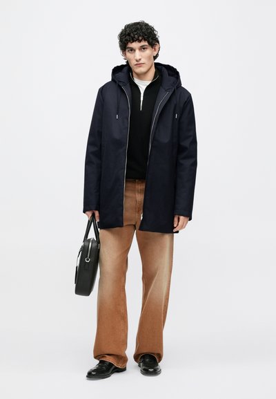 Tumesinine kapuutsiga mantel lukuga ees, must kampsun ja helepruunid lahenevate sääreosadega teksad. Käes must kott, seismas ühevärvilise tausta ees.