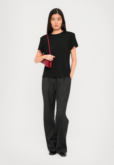 Woman wearing a black t-shirt and loose black pants, holding a small red shoulder bag, standing against a plain light background.