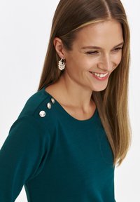 Niebieskozielona bluzka z długim rękawem i okrągłym dekoltem, zdobiona trzema dekoracyjnymi guzikami na lewym ramieniu; gładki materiał o dopasowanym kroju.