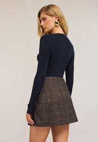 Kvinna med blont hår som bär stora guldörhängen, en marinblå rippad långärmad topp och en brun rutig minikjol, vänd åt sidan.