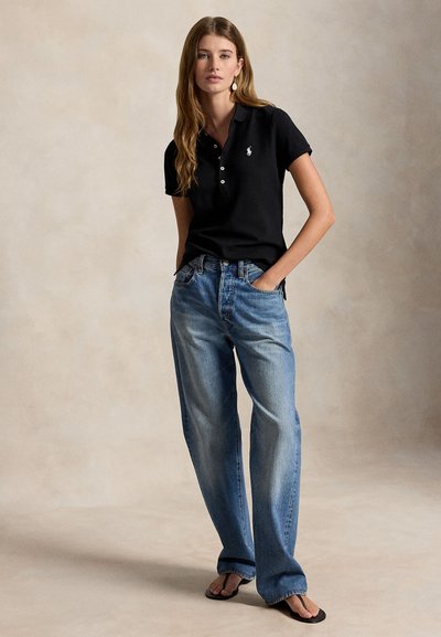 Γυναίκα όρθια με τα χέρια στις τσέπες, φοράει μαύρο πόλο κοντομάνικο, άνετο μπλε τζιν και μαύρα σαγιονάρες σε ομοιόμορφο φόντο.