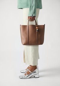 Braune Ledertasche mit strukturiertem Finish, ausgestattet mit zwei vertikalen Nähten und bunten Perlenakzenten am Griff. Silbere Hurensandalen mit Riemen.