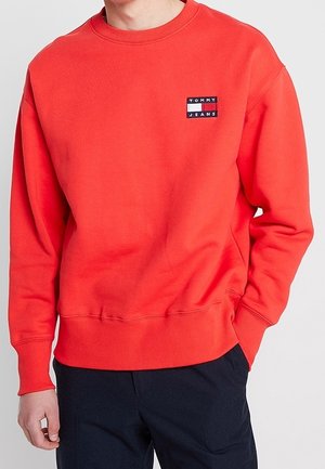 Mężczyzna w jaskrawoczerwonej bluzie Tommy Jeans z prążkowanymi mankietami i dołem, w połączeniu z ciemnymi spodniami, stoi z jedną ręką w kieszeni.