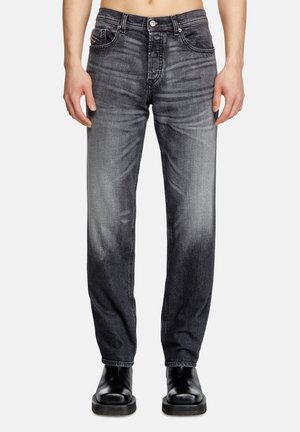 Mand iført falmede sorte jeans med lige ben og sorte læderstøvler, stående med armene ned langs siden mod en ensfarvet hvid baggrund.