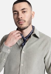 Cămașă verde deschis cu dungi, cu guler butonat, mâneci lungi și o eșarfă cu model bleumarin legată la gât. Textură netedă.