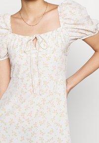 Robe florale en tissu léger avec un décolleté carré, des manches bouffantes et un détail à nouer sur le devant ; arbore un motif de petites fleurs roses et de feuillage.