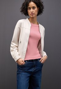 Lichtbeige bomberjack met elastische manchetten, gedragen over een zachte roze shirt. Het model draagt ook donkerblauwe jeans, met de handen in de zakken.