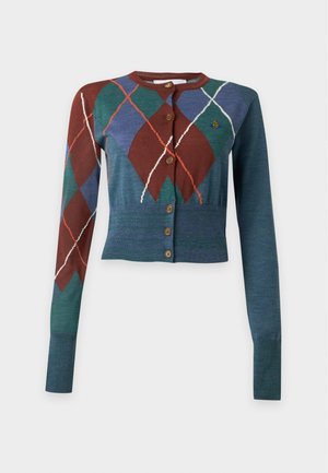 Joven mujer modelando un cárdigan argyle multicolor sobre una camisa blanca, falda de cuero burdeos y zapatos slingback negros con punta, de pie frente a un fondo blanco.