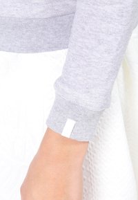 Cuff di un maglione grigio con lavorazione a maglia testurizzata. Presenta un'etichetta bianca sul lato esterno. Lo sfondo mostra un tessuto bianco con motivo.