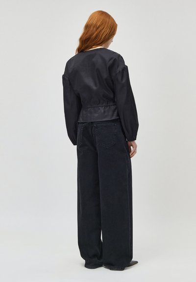 Femme rousse de dos, portant un chemisier noir à manches longues avec taille froncée et un jean noir à jambes larges, debout sur un fond uni.