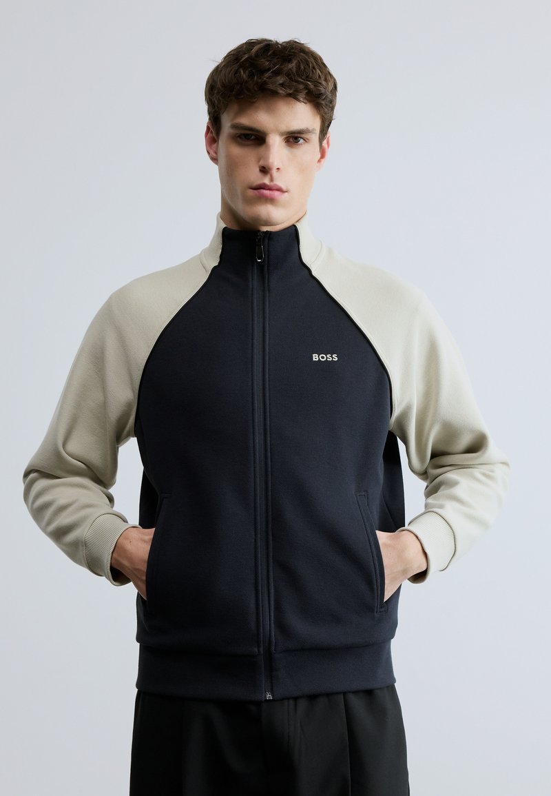 Sudadera con cremallera en azul marino y beige con cuello alto, mangas raglán y bolsillos laterales; con logo en el pecho izquierdo.