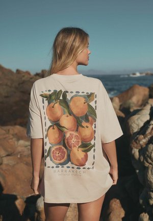 Woman standing on rocky shore wearing beige shirt with orange fruit design and "Marrakech Mandarin Oranges" text on the back.