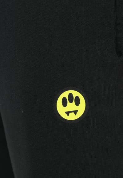 Schwarzer Baumwollstoff mit einem gelben, kreisförmigen Grafikdesign, das drei Punkte und Zähne zeigt, platziert an der Seite des Kleidungsstücks.