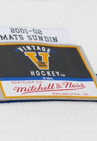 Etichetta in tessuto bianca con il testo "2001-02 MATS SUNDIN", un rettangolo nero con un logo "V" giallo e "Mitchell & Ness" in rosso.