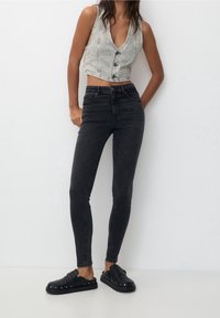 Femme portant un gilet en jean gris sans manches boutonné, un jean skinny noir taille haute et des chaussures noires à enfiler ornées de clous, debout contre un mur blanc.