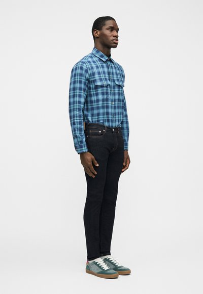 Άνδρας που φοράει μπλε και πράσινο πουκάμισο καρό, σκούρο στενό τζιν και παπούτσια τυρκουάζ, που στέκεται μπροστά σε απλό λευκό φόντο.