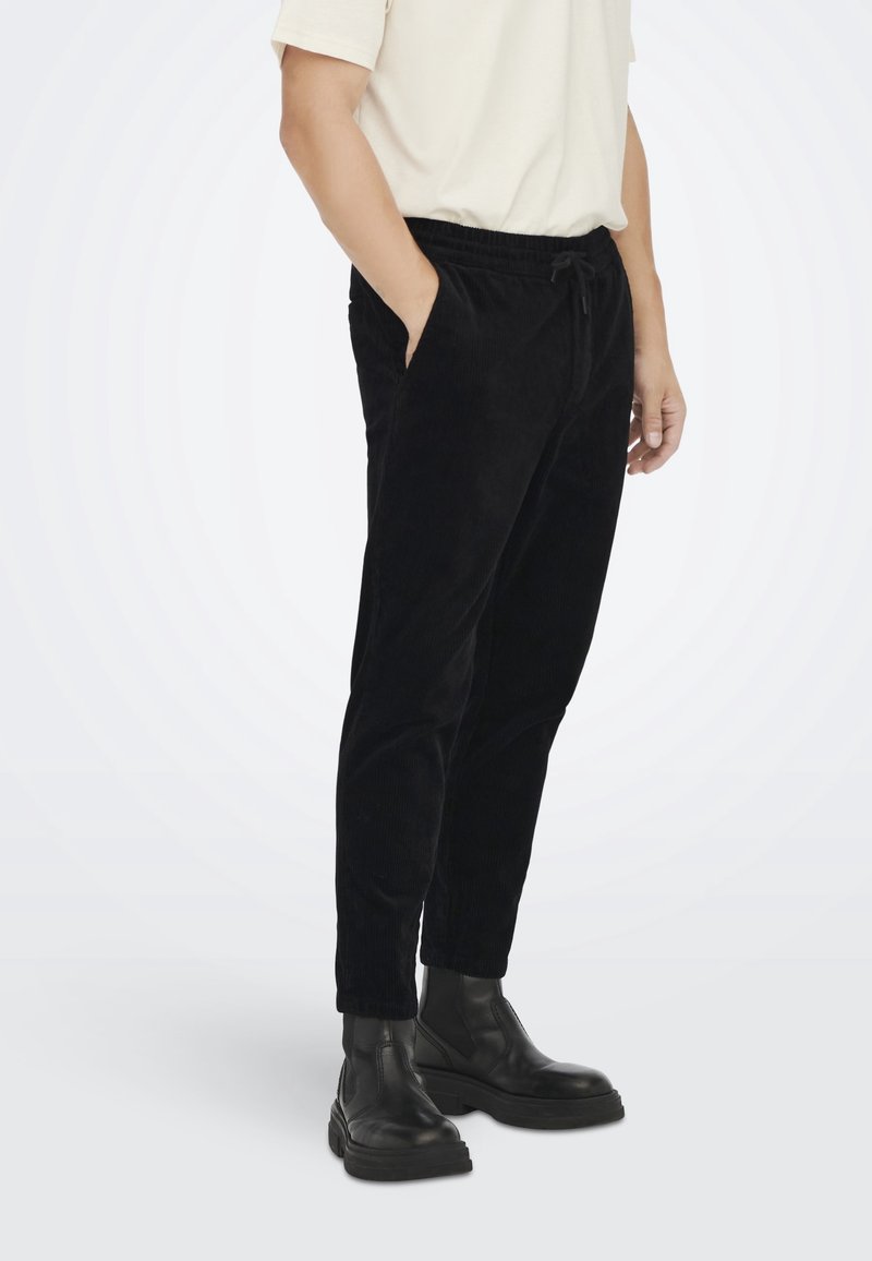 Personne portant un pantalon en velours côtelé noir avec une taille élastique et des bottines en cuir noir, mains partiellement dans les poches.