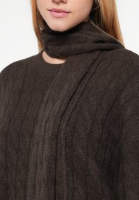 Brun stickad tröja med texturerat flätmönster, ribbad rund halsringning och draperat tyg runt halsen för extra värme.