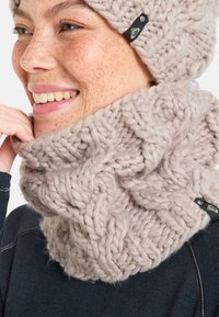 Gehaakte sjaal in zacht beige met een chunky textuur, met een gevlochten patroon en een zwarte label met logo. Sjaal is om de nek gewikkeld.
