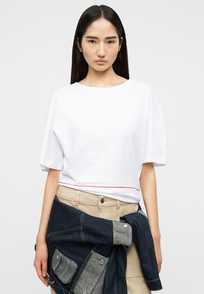 Νεαρή γυναίκα με μακριά σκούρα μαλλιά φοράει λευκό κοντομάνικο τοπ και μπεζ παντελόνι, κρατώντας ένα διπλωμένο τζιν μπουφάν γύρω από τη μέση της.