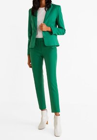 Costume vert comprenant un blazer ajusté et un pantalon sur mesure, en tissu lisse. Associé à des bottines blanches pour un contraste.
