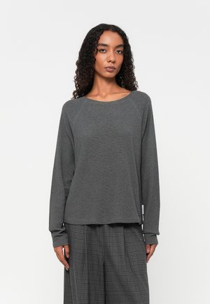 Maglietta grigia a maniche lunghe con scollo rotondo, realizzata in tessuto morbido. Presenta maniche raglan e una vestibilità comoda. Indossata con pantaloni grigi a quadri.