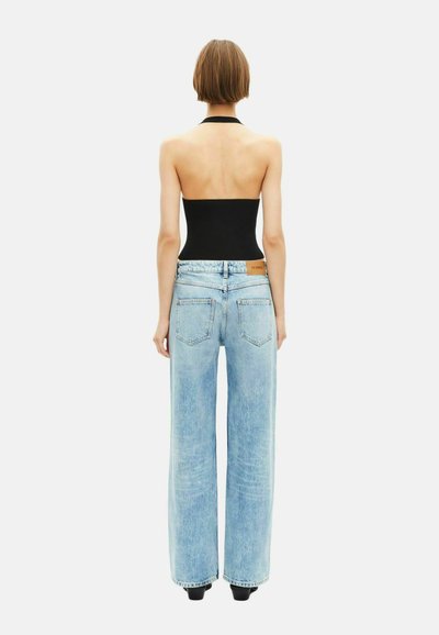 Vrouw met kort haar draagt een zwarte haltertop en lichtblauwe hoge wijde jeans, staande met de rug naar de camera op een witte achtergrond.