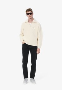Suéter de punto texturizado en crema con media cremallera y cuello ancho, combinado con pantalones negros ajustados y zapatillas grises, completado con gafas de sol oscuras.
