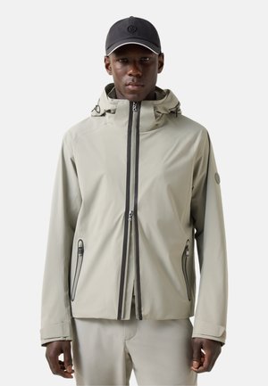 Mand iført let beige hættetrøje med sorte lynlåse og lommer, kombineret med matchende bukser og en sort kasket.