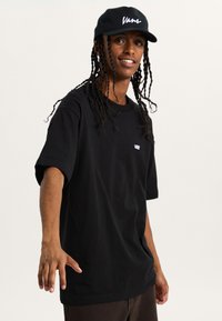 Schwarzes T-Shirt mit kurzen Ärmeln und Vans-Logo auf der linken Brust, kombiniert mit einer schwarzen Baseballmütze, auf der der Vans-Schriftzug in Weiß zu sehen ist.