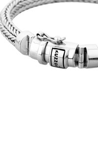 Sudraba pītā aproce ar metālisku aizdari, uz kuras ir logo zīme. Virsma ir gluda ar spīdīgiem akcentiem un pulētu apdari.