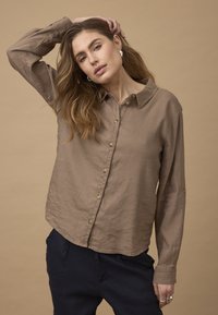 Jeune femme aux longs cheveux ondulés portant une chemise marron boutonnée et un pantalon foncé, posant avec une main sur la tête devant un fond neutre.