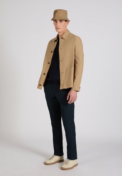 Uomo che indossa un cappello a tesa larga color sabbia, giacca beige con bottoni, maglietta nera, pantaloni scuri e scarpe da ginnastica color crema, in piedi contro uno sfondo chiaro e uniforme.