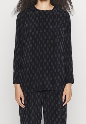 Femme portant un haut noir à manches longues et un pantalon avec un motif à carreaux sombre subtil, debout devant un fond clair uni.