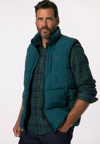 Teal puffer vest met zwarte accenten, ritszakken en een logo. Draag het over een groene en marineblauwe geruite flanellen-shirt met lange mouwen.