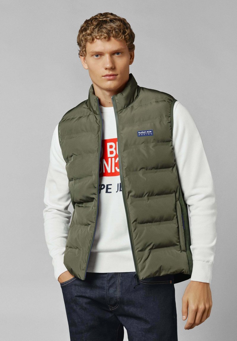 Groene gewatteerde vest met ritssluiting, hoge kraag en logo-detail. Draagt over een witte trui met rode en marineblauwe tekst. Donkere spijkerbroek zichtbaar.
