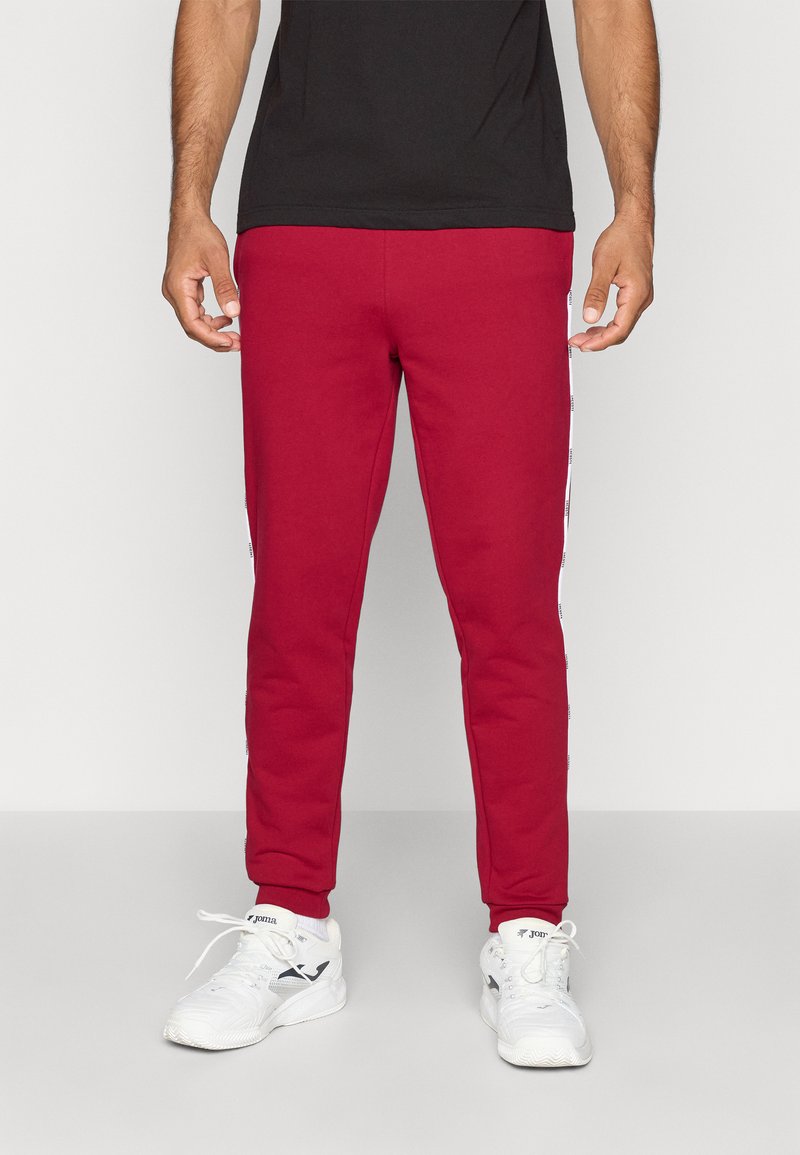 Red sweatpants with a slim fit, made of soft fabric, featuring white side stripes with text. Worn with white athletic shoes.