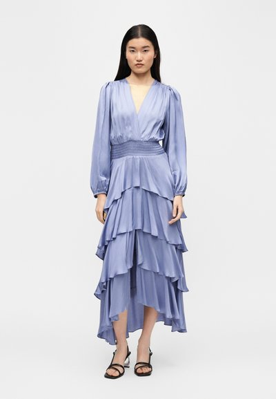 Γυναίκα που στέκεται φορώντας ένα ανοιχτό μωβ φόρεμα με στρώσεις, μακρυά φουσκωτά μανίκια και μαύρα ανοιχτά πέδιλα σε απλό λευκό φόντο.