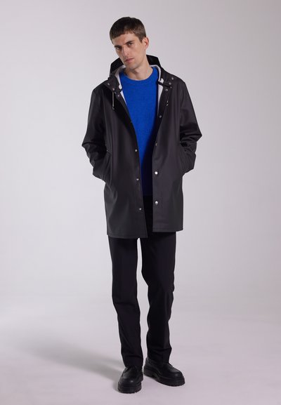 Svart vattentät kappa med silverfärgade tryckknappar, huva och sidofickor; bärs över en blå tröja och svarta byxor. Svarta skor med grov sula.